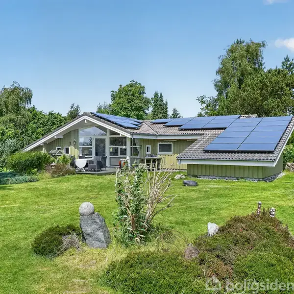Grønt sommerhus med terrasse; bord og stole står i solen. Omgivet af frodig have og træer.