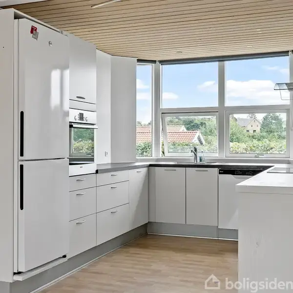 Et moderne, rummeligt køkken med hvide skabe og indbygningsapparater. Rummet er lyst med et stort vindue ud mod en have. Trægulv og træbeklædt loft skaber en varm atmosfære.