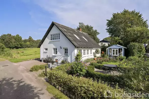Huset er hvidt med stråtækt tag, omgivet af en have med havemøbler. Det ligger ved en vej i en landlig omgivelse med marker og træer i baggrunden.