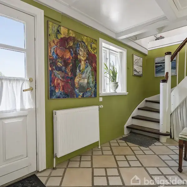 En hvid dør med vinduer står i et farverigt rum med grønne vægge. Ved siden er en rektangulær radiator, ved siden af et farverigt maleri. Trappe fører opad.