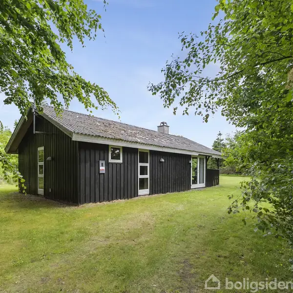 Et sort træhus står omgivet af grønne træer og græsplæne i solrigt vejr. Huset har store vinduer og en skorstenspibe. En diskret skiltning hænger ved indgangen.