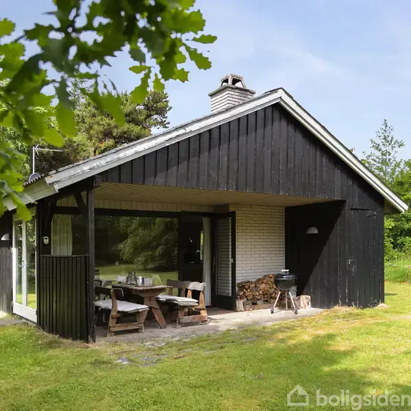 Et sortmalet sommerhus står stille i en frodig have. Et overdækket terrasseområde har et træbord og bænk. Omgivet af græs og tætte grønne træer.