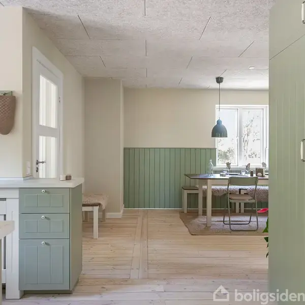 Køkken med grønne skabe har åbning til spiseområde. Spisebordet står ved vindue, omgivet af stole og belysning ovenfra. Lyst trægulv. Ingen tekst bortset fra "boligsiden".