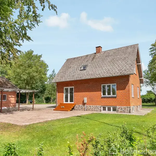 Et rødt murstenshus med skråt tag står på en græsplæne omgivet af træer. Huset har en stor terrasse foran og flere vinduer.