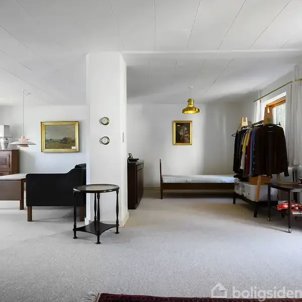 Et stort, lyst værelse indeholder en seng, en sofa og en tøjstativ. Væggene prydes med kunst. Rummet har store vinduer med gardiner, der giver et luftigt helhedsindtryk.