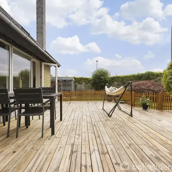 En træterrasse med bord og stole til venstre står ved et hus. En hængekøje er placeret til højre. Haven er omgivet af et hegn og grøn beplantning.