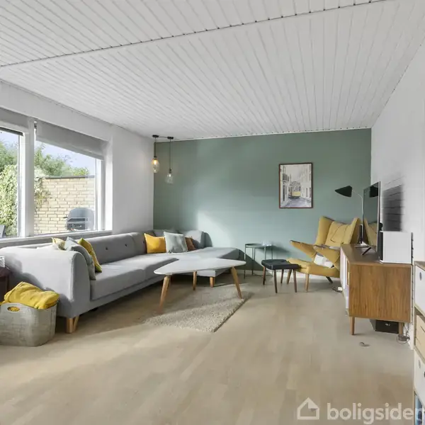 En stue med en grå sofa og to lænestole. Et sofabord foran sofaen. På væggen hænger et maleri. Store vinduer viser en have udenfor. En skænk med flasker til højre.