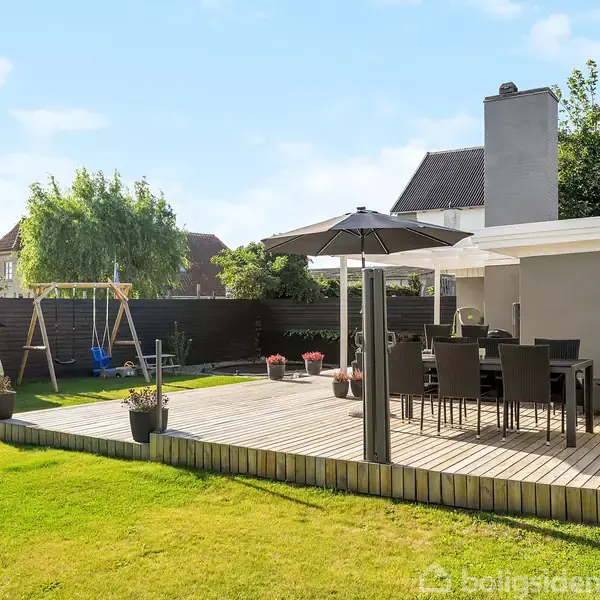 Terrasse med havemøbler og parasol, placeret i en velplejet have. I baggrunden ses en gynge og skur, omgivet af grøn græsplæne og hække under en klar blå himmel.