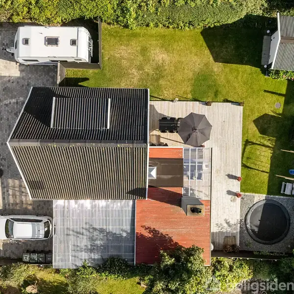 Et hus set oppefra med en stor terrasse, en parasol, og en trampolin i haven. En bil og en campingvogn står på indkørslen. Omkring huset er grøn vegetation.