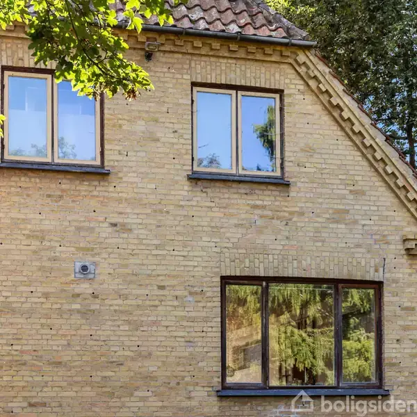 Et gult murstenshus står stille med tre vinduer, omgivet af frodig vegetation og store træer under en blå himmel.