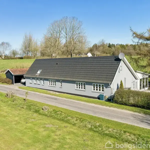 Hus med gråt tag står langs en vej omkranset af grønne marker og træer. I nærheden er en lille garage med rødt tag, omkranset af åben natur og blå himmel.