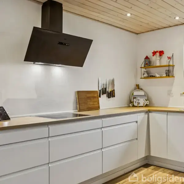 Et moderne køkken med hvide skabe og træbordplader. En sort emhætte hænger over komfuret, og der er knive på væggen. Hylder med blomster og planter ved vinduet.