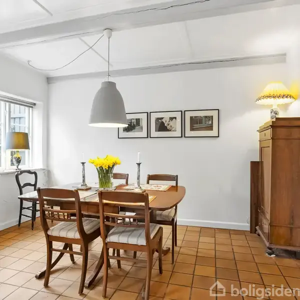Spisebord med seks stole står i et lyst rum. Vase med gule blomster på bordet. Væghængte billeder, skænk, og en lampe skaber hyggelig atmosfære i rummet med terracotta-gulv.