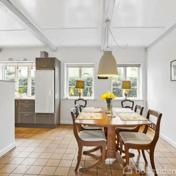 Spisebord med blomster i vase omgivet af stole står i lyst køkken-alrum med hvide vægge, fliser på gulvet, og billeder på væggen. Sollys strømmer ind gennem vinduerne.