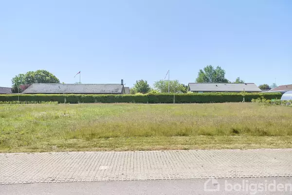 En tom byggegrund ligger stille i forgrunden omgivet af græs. I baggrunden ses en række huse med hække og træer under en klar blå himmel.