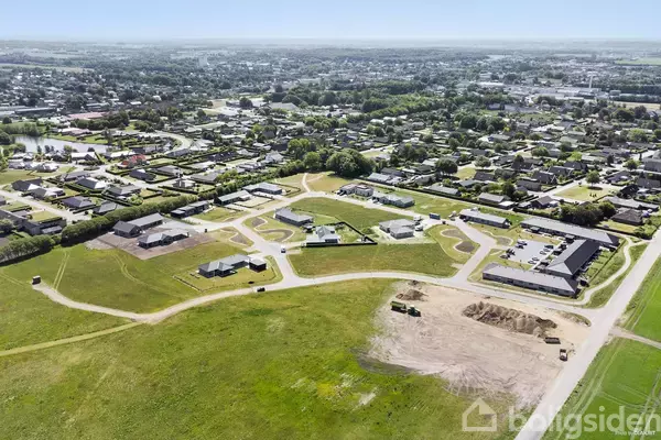 Luftfoto viser en bydel med rækkehuse, spredt efter veje, omgivet af grønne områder. I forgrunden ses åbent græsland og jord, muligvis forberedt til byggeri.