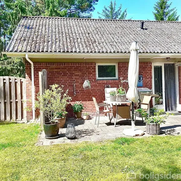 Et rødt murstenshus med fladt tag har en terrasse med havemøbler og planter. Terrassen er omgivet af grøn græsplæne og skov. Billedet indeholder ingen tekst.