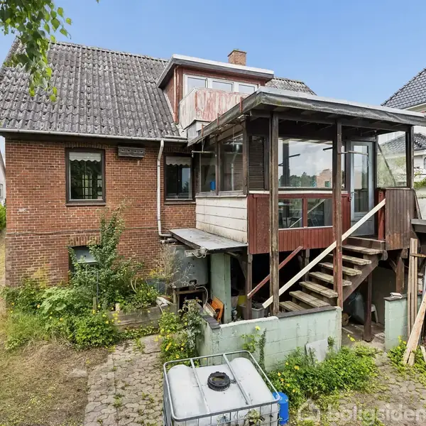 Et rødt murstenshus med en tilbygning i træ ses bagfra med en trappe op til en lukket veranda. Omkringliggende have er delvist tilgroet med græs og buske.