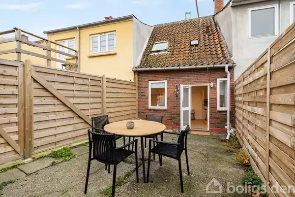 Rundt havebord med fire stole står på en flisebelagt terrasse. En mindre murstensbygning med tagvinduer er i baggrunden. Terrasse er omgivet af træhegn.