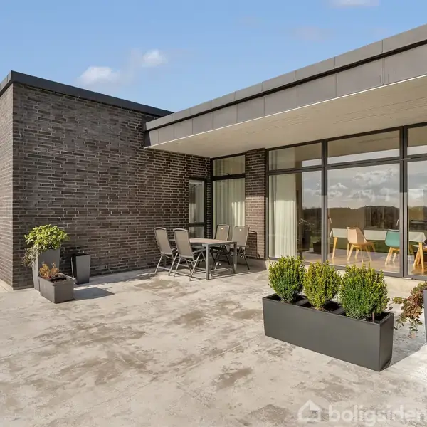 En moderne murstensbygning med store vinduespartier. Et udemøblement står på terrassen med plantekasser. Omkringliggende græsplæne og blå himmel skaber en lys og åben atmosfære.
