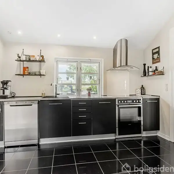 Sort køkken med fliser på gulvet og sort-hvide skabe; opvaskemaskine og ovn ved siden af hinanden. To hylder med køkkenting over, vindue i midten.