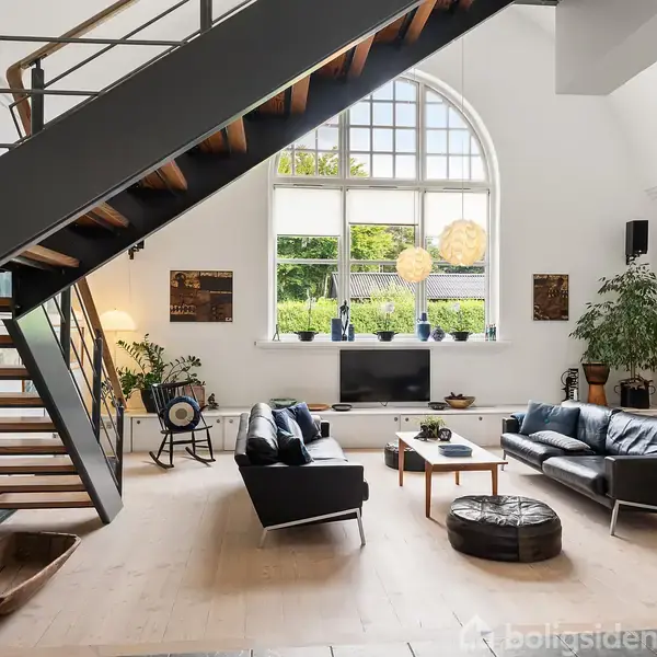 Stue med moderne møbler, hvor to sorte sofaer er placeret omkring et sofabord. Ovenover er en trappe. Store vinduer giver lysindfald. Grønne planter og billeder pryder rummet.