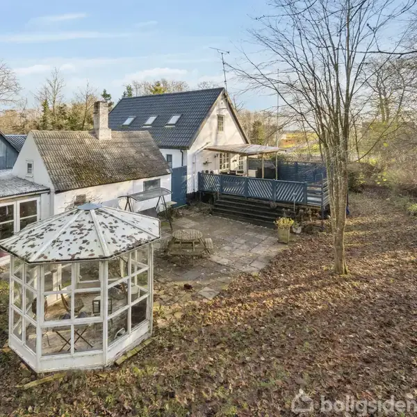 En hvid havepavillon står stille på en græsplæne omgivet af en stor ejendom med flere bygninger og et terrasseområde. Træer omkranser området under en blå himmel.