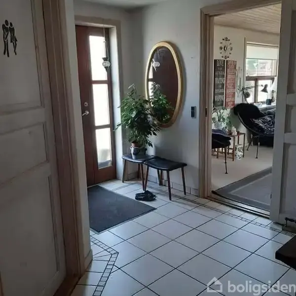 En gang med flisegulv og en dør med glaspanel. En plante står på en bænk under et ovalt spejl på væggen. Indtil stuen ses møbler og dekorationer.