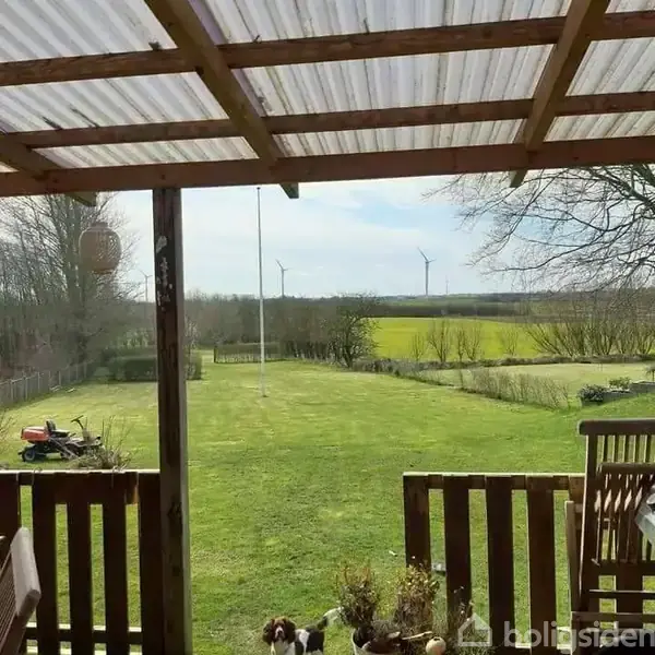 Terrasse med trætag kigger ud over en velholdt græsplæne med flagstang. I baggrunden ses marker og vindmøller under en klar himmel. En plæneklipper står til venstre.