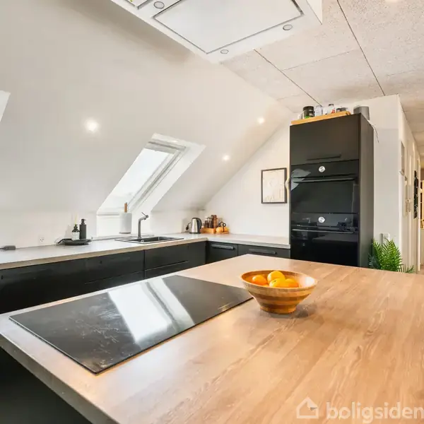 Moderne køkken med sort indretning og skråvinduer. En skål med appelsiner står på en træbordplade i et lyst rum med hvide vægge og lofter. Korridor i baggrunden.