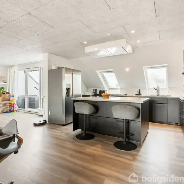 Køkken med central ø, sorte skabe og barstole. Store vinduer giver naturligt lys. I baggrunden ses åbne hylder med dekorationer og en stue med legetøj. Plante i hjørnet.
