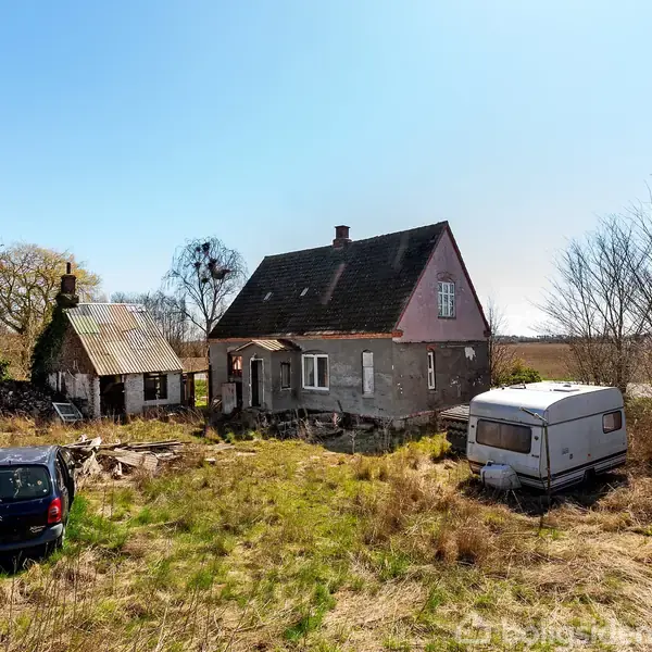 Et forladt hus står midt på en tilgroet grund. Ved siden af huset ses en gammel bil og en campingvogn. Omkringliggende træer og buske tilføjer til det øde landskab.