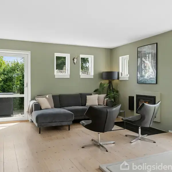 Sofa i vinkel med puder, står ved siden af to sorte stole i en moderne stue med trægulv, grønne vægge, store vinduer, pejs og dør, der fører ud til have.