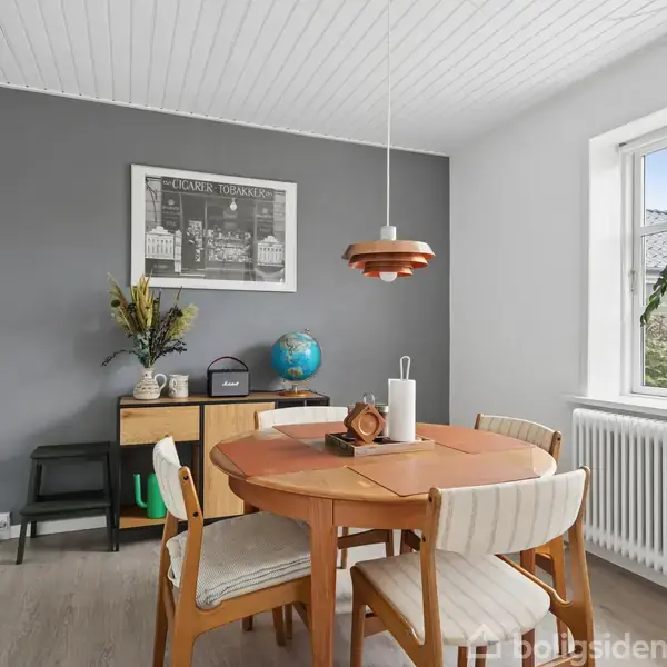 Rundt spisebord med fire stole står i et lyst rum. På væggen hænger et billede med teksten "CLARK TOWERS," og et lille skab står i hjørnet med en globus og planter.