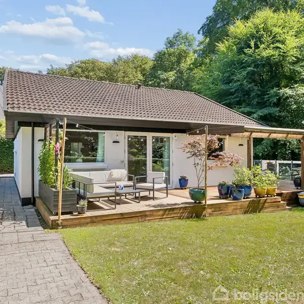 Et lille hus med en træterrasse, hvorpå der er havemøbler. Omgivet af en velplejet græsplæne og grønne træer i baggrunden, under en klar, blå himmel.
