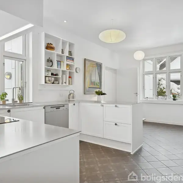 Et moderne køkken med hvide skabe og hængende lamper. Der står små planter i vindueskarmen. Rummet er lyst med store vinduer og har grå flisegulv. Ingen tekst er synlig.