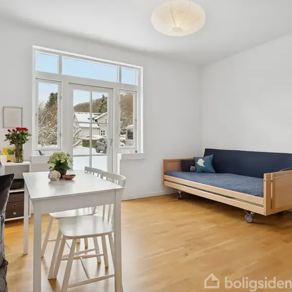 En lys dagligstue har en sovesofa med blå madras ved væggen, et hvidt bord med stole foran et stort vindue, og trægulv danner rammen.