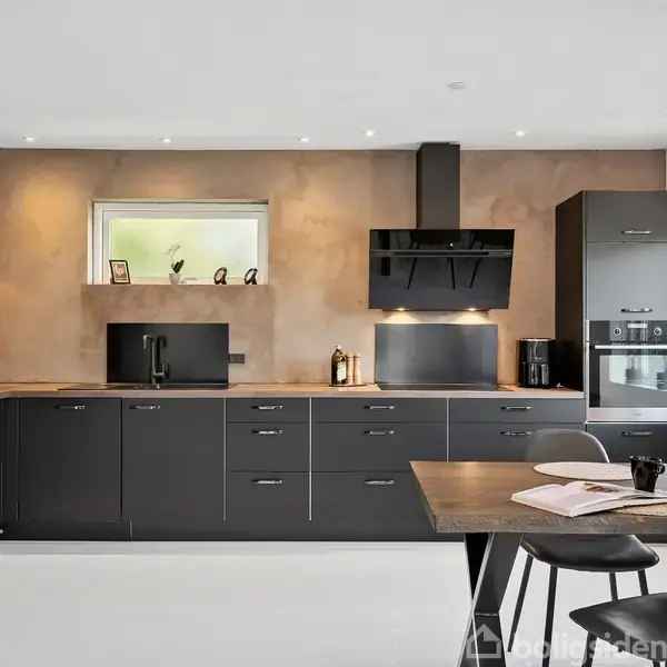 Et moderne køkken med sorte skabe og skuffer. Overbordet hænger en industriel lampe. Et spisebord med stole står til højre. Vinduet til venstre giver lys til rummet.