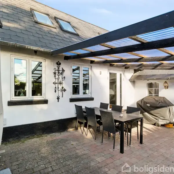 En udendørs terrasse med et sort spisebord og seks stole under en pergola. En grill er ved siden af en glasdør. Terrasse fliser og hvide vægge omgiver området.