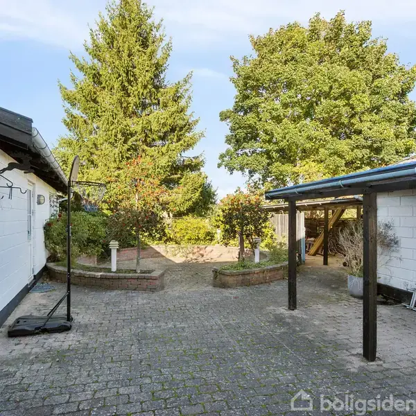Terrasse med et sort spisebord og seks stole under en transparent overdækning. Til venstre står en sammenfoldet parasol. Ved siden af står en grill dækket med et sort betræk.