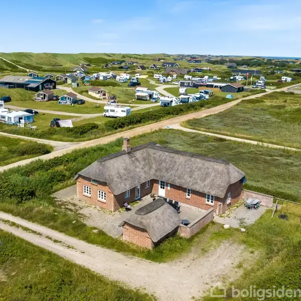 Et stråtækt hus ligger stille i et åbent, grønligt landskab med snoede veje. I baggrunden ses spredte huse og campingvogne i en sommerhusområde.