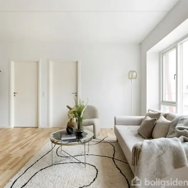 En lys stue med sofa og tæppe; sofaen står til højre foran et stort vindue. En plante og to døre er til venstre. Stuebordet har en dekorativ vase.