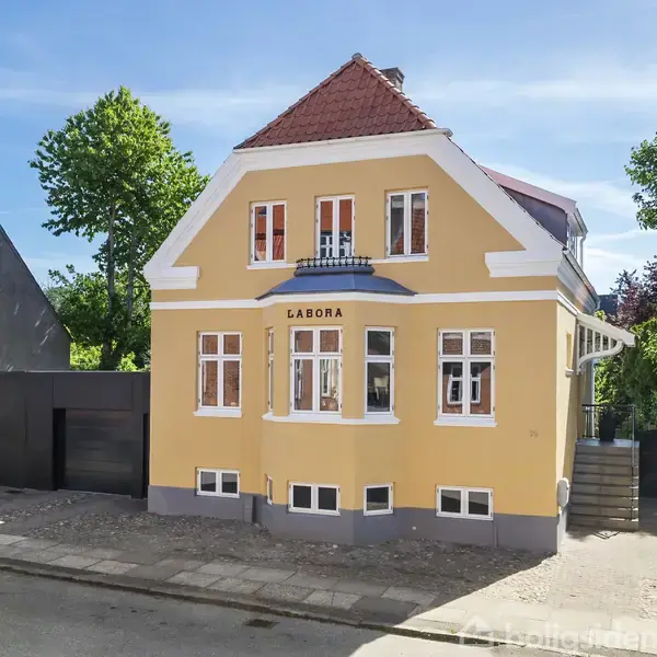 Gul bygning med rødt tag står på en vej. Tekst “LABORA” er placeret over hoveddøren. Bil er parkeret til højre, træer og klare blå himmel omkranser huset.