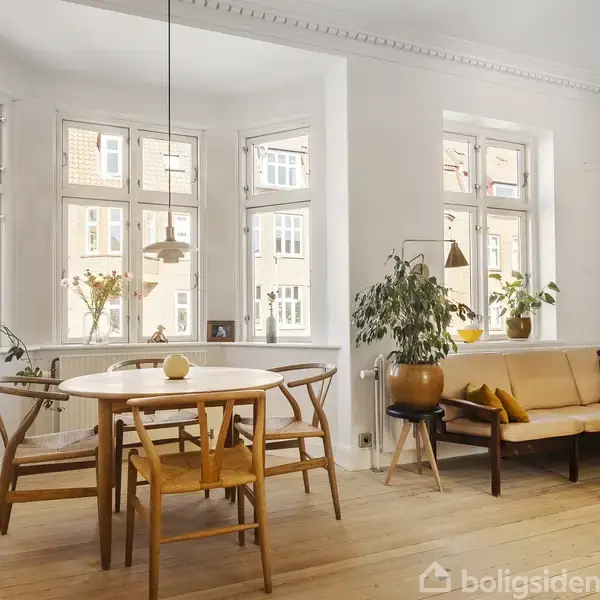 Det lyse rum indeholder et rundt træbord med stole, omkranset af store vinduer. Ved siden af står en sofa med puder og planter i vindueskarmen, der skaber en hyggelig atmosfære.