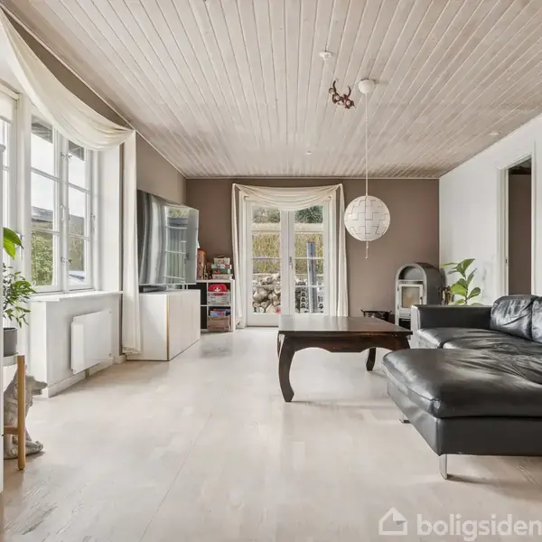 Sort læderhjørnesofa står i en rummelig stue med lyse trægulve. Store vinduer og glasdøre slipper naturligt lys ind, mens væg til loft-skabe og planter skaber en hyggelig atmosfære.