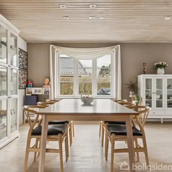 Et rektangulært spisebord med seks stole står i en lys spisestue. Store skabe langs væggene indeholder udstillinger. Et stort vindue i baggrunden giver udsigt til en hæk.