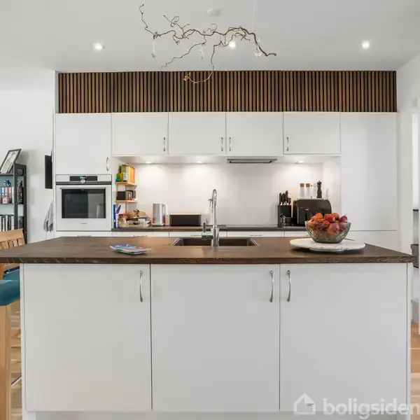 En hvid køkkenø med vask står i centrum, omgivet af skabe, ovn og oplyste hylder. Baggrunden viser en spisestue med stole, bøger og dekorationer.