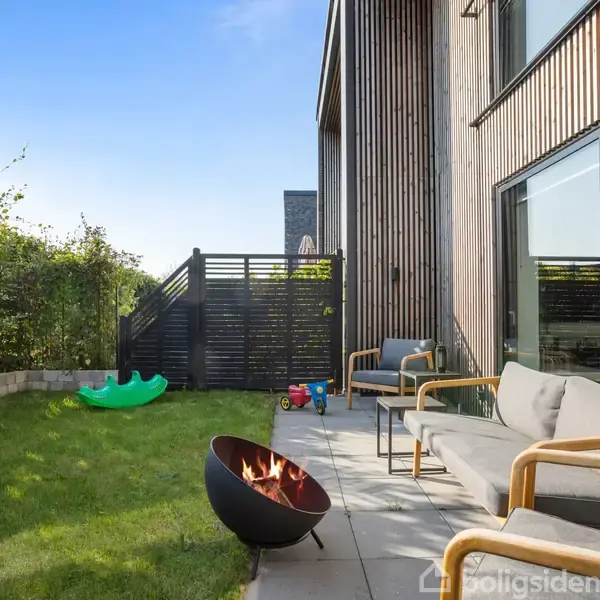 En moderne terrasse med to sofaer og en ildskål, hvor ilden brænder. Græsplæne med legetøj i forgrunden, omgivet af en hæk og et trægærde.