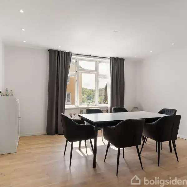 Et spisebord med seks sorte stole står i et lyst rum med trægulv. Store vinduer med mørke gardiner slipper naturligt lys ind fra venstre.