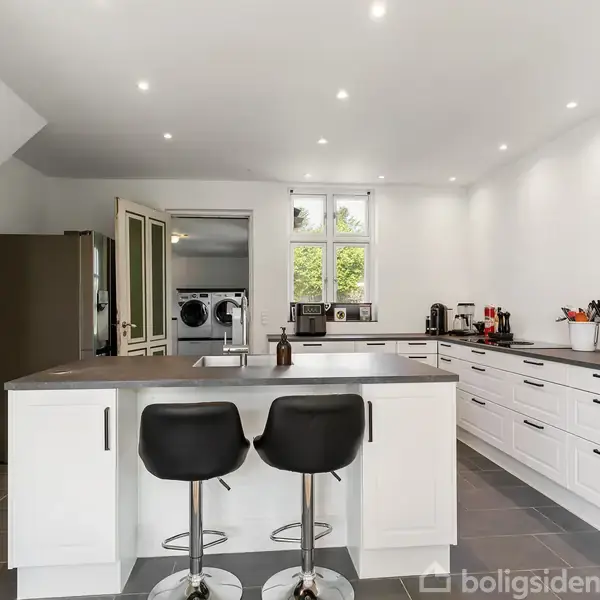 Et moderne køkken med en stor ø i midten, som har to sorte barstole. Hvide skabe og skuffer omgiver rummet; en vaskemaskine er synlig i baggrunden ved vinduet.